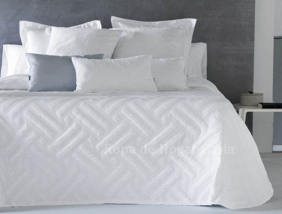 Cama con edredón blanco y varios cojines en tonos claros sobre fondo gris.