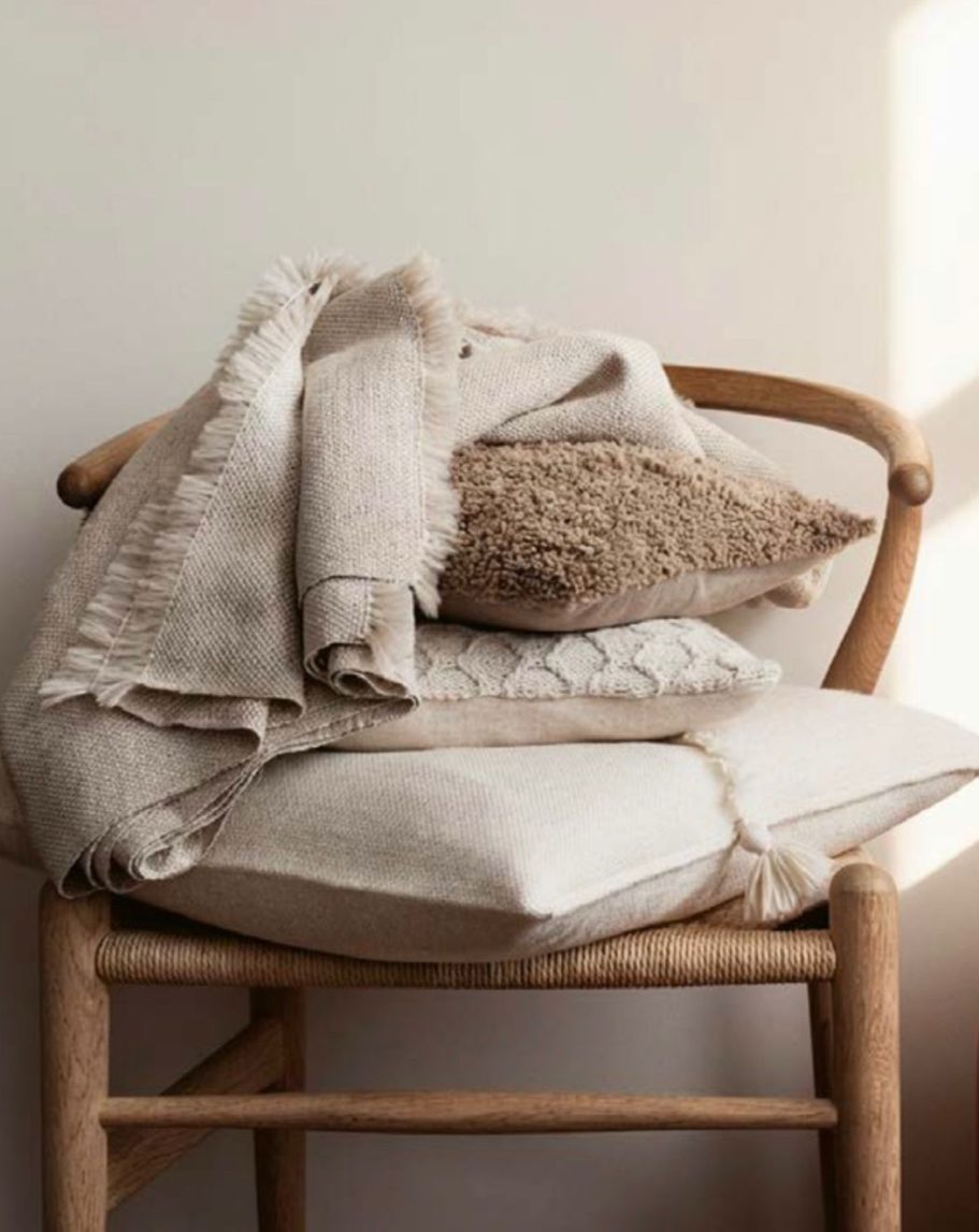 Silla de madera con cojines y mantas en tonos neutros apilados.