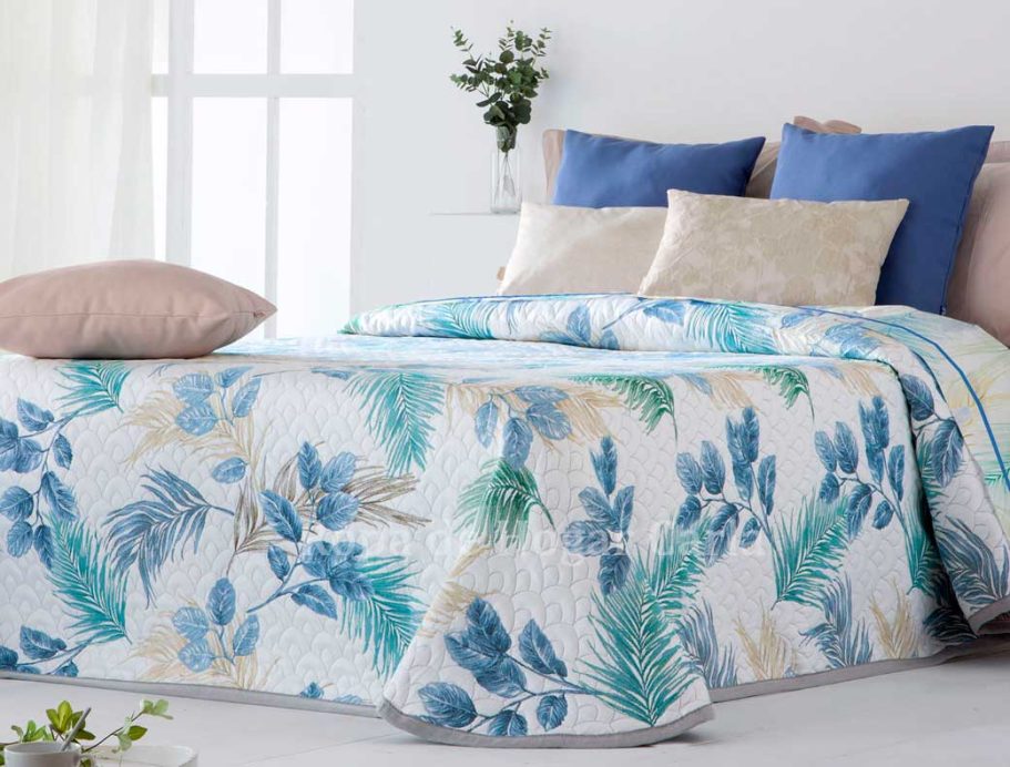 Colcha con diseño de hojas en tonos azules y verdes sobre una cama con cojines decorativos.