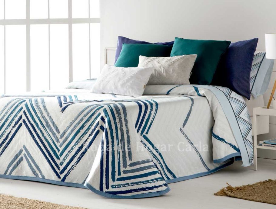 Cama con colcha de diseño geométrico en tonos blancos y azules, almohadas decorativas.